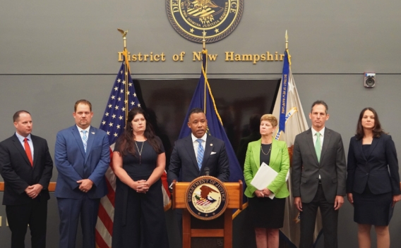 Assistant Attorney General Kenneth A. Polite, Jr. speaks at the podium with a Department of Justice seal. The background displays the seal of the district of New Hampshire.