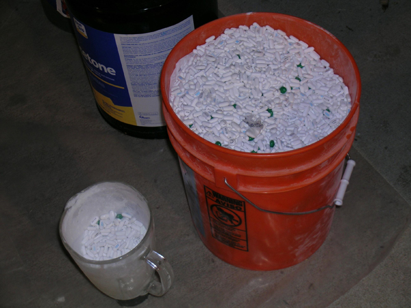 Photo of a 5-gallon bucket of pills, a glass measuring cup with pills, and a 5-gallon can of acetone.