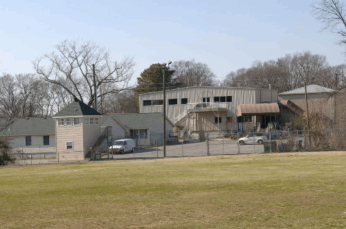 Photograph of a fenced in property featuring a two-story, 12 x 12 foot steel guard shack with a green roof situated on the left of this photo. The photo also shows two larger steel buildings, and a parking lot with two cars.