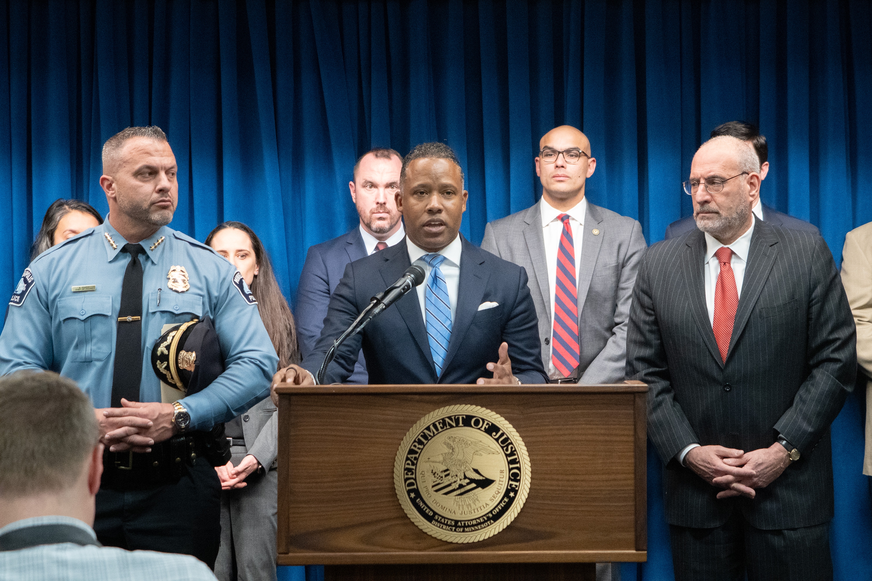 Assistant Attorney General of the Criminal Divison Kenneth A. Polite, Jr. delivers remarks from a podium bearing the United States Attorney's Office for the District of Minnesota seal. To the left stands the Chief of the Minneapolis Police Department Brian O'Hara. To the right stands United States Attorney for the District of Minnesota Andrew M. Luger. Other officials stand in a row behind.