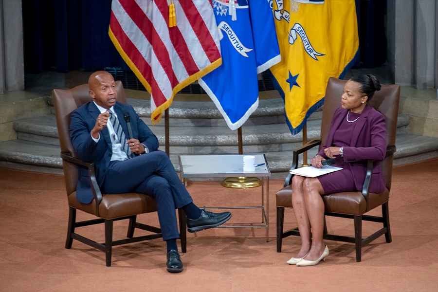Assistant Attorney General for Civil Rights Kristen Clarke and Equal Justice Initiative Executive Director Bryan Stevenson participate in a seated discussion in the Great Hall at the Department of Justice. Behind them are the flags of the United States and The Justice Department.