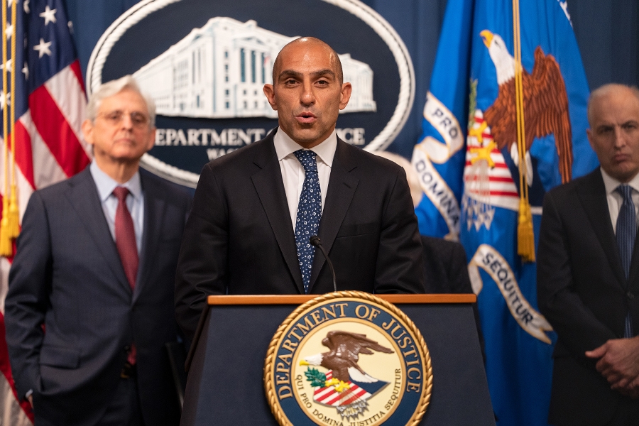 Chairman Rostin Behnam of the Commodity Futures Trading Commission (CFTC) delivers remarks from a podium at the Department of Justice. To the left is Attorney General Merrick B. Garland, and to the right is Assistant Attorney General for National Security Matthew G. Olsen.
