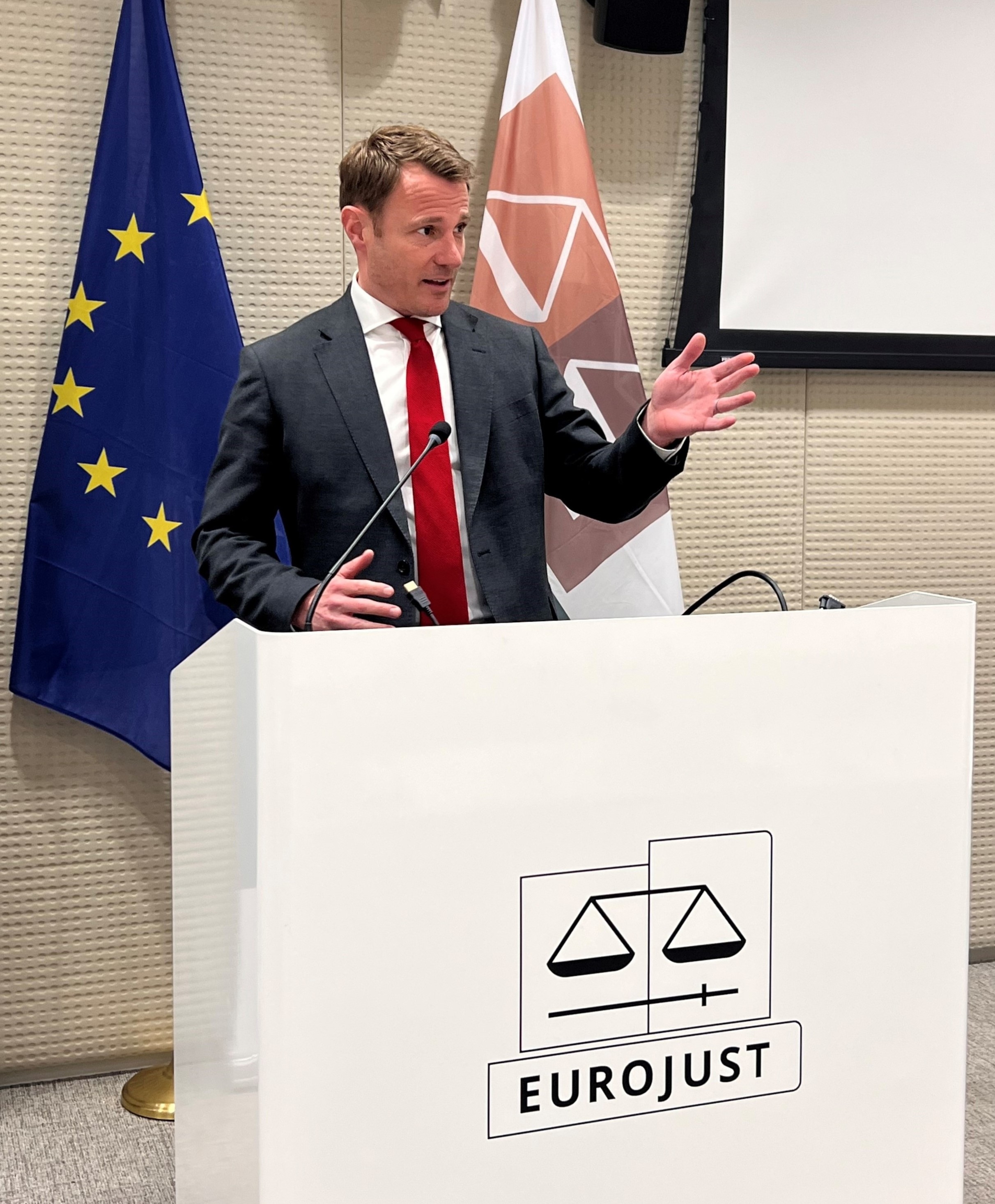 A person standing at a podium with a microphone and flags behind him.
