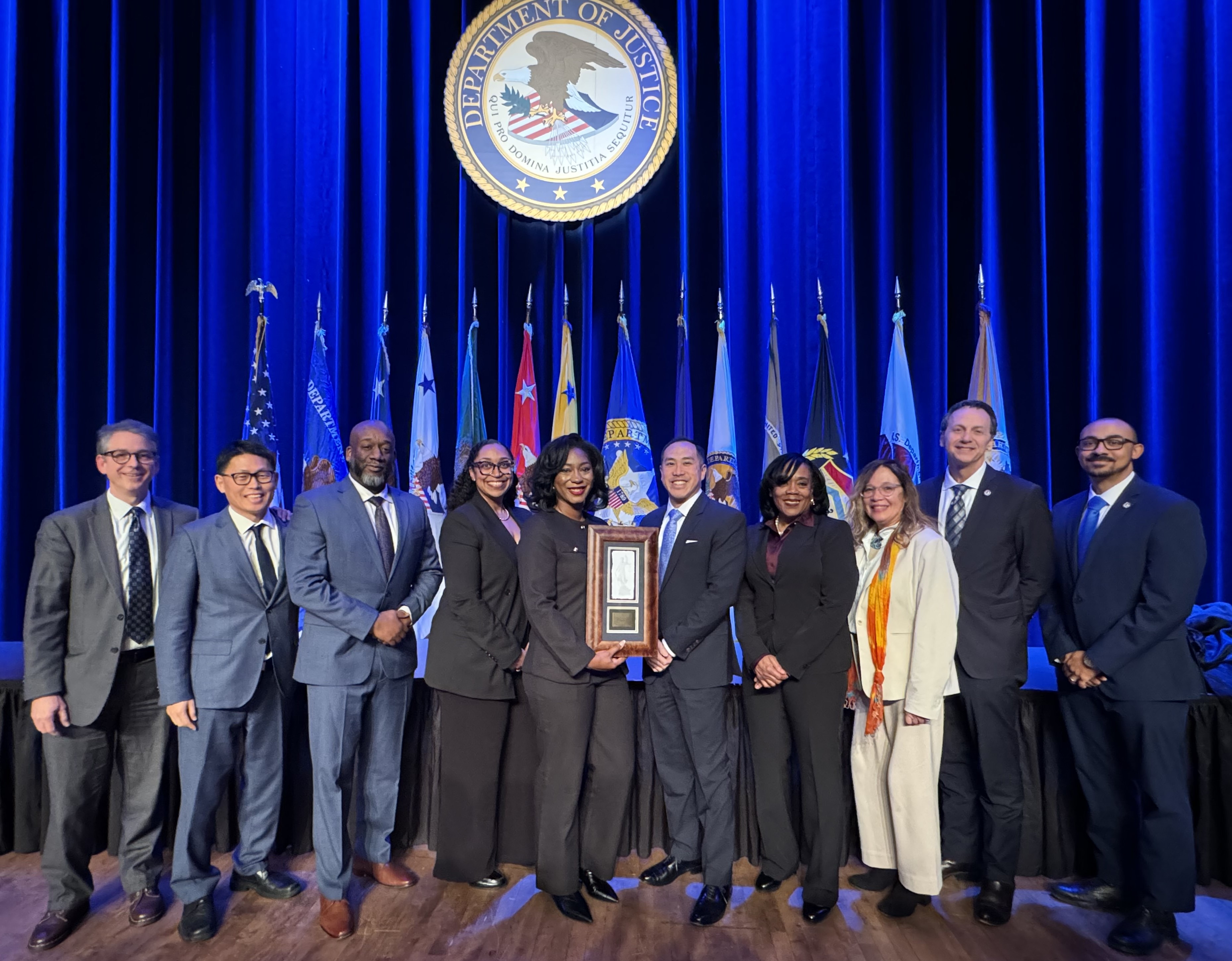 10 people standing on stage, under DOJ Seal, holding 1 plaque, posing for photo