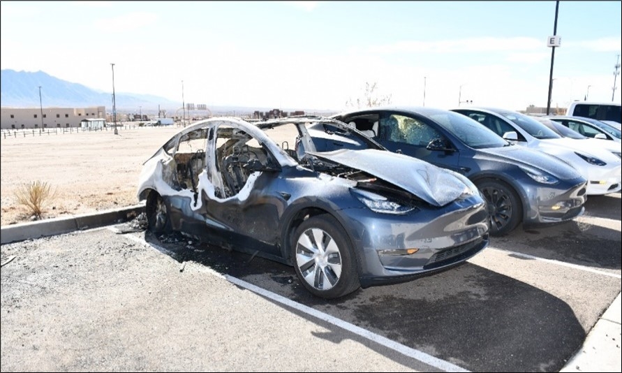 A front-profile view of a Tesla parked outside, with doors, hood, and windshield severely damaged.