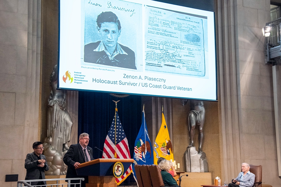 U.S. Department of Veteran’s Affairs employee Ronald Zenon Piaseczny delivers remarks from a podium in the Great Hall at the Department of Justice. On a projection screen behind him is an image of his father Zenon A. Piaseczny and his identification card from Poland in 1945.