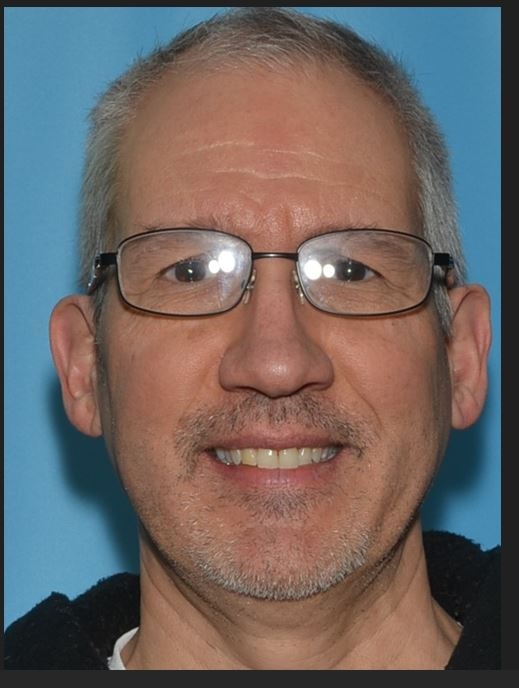 Man with grey hair and glasses against blue wall.