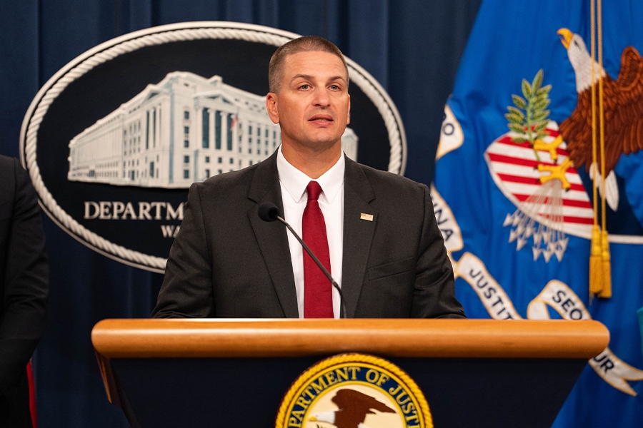 Christopher Delzotto delivers remarks from a podium at the Department of Justice. He is joined by Department of Justice and federal government officials.