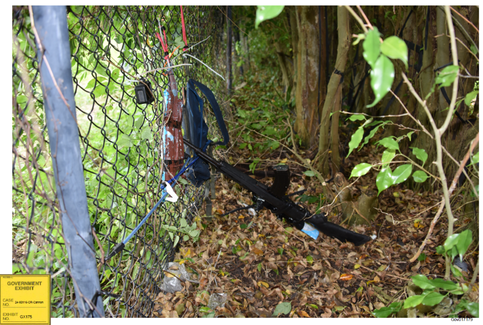 Black rifle on the ground and pointing through brush and chain-link fence.