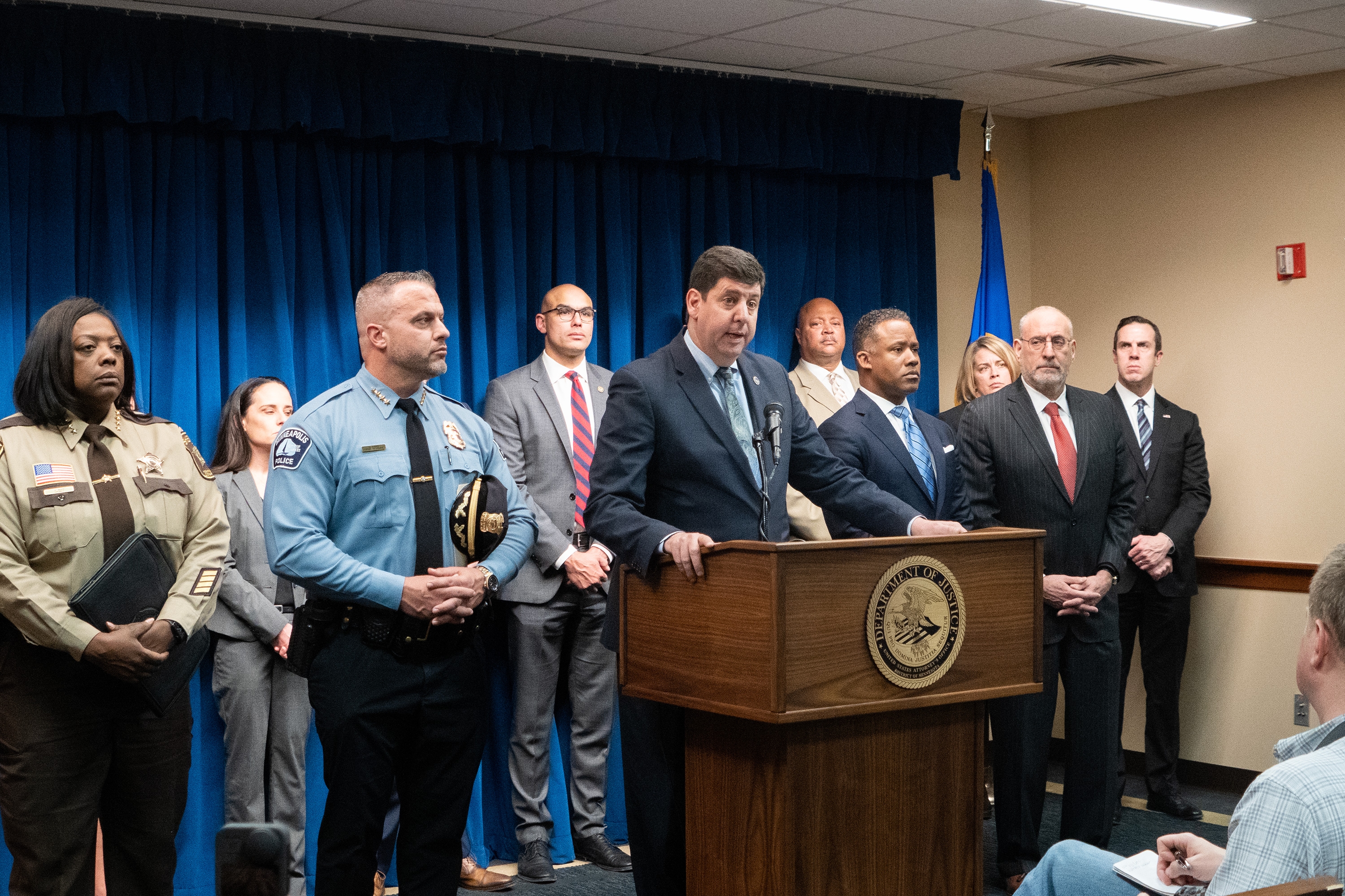 ATF Director Steven M. Dettelbach delivers remarks from a podium bearing the United States Attorney's Office for the District of Minnesota seal. To the left stands Chief of the Minneapolis Police Department Brian O'Hara (left) and Hennepin County Sheriff Dawanna S. Witt (far left). To the right stands Assistant Attorney General Kenneth A. Polite, Jr. (right) and United States Attorney for the District of Minnesota Andrew M. Luger (far right). Other officials stand in a row behind.