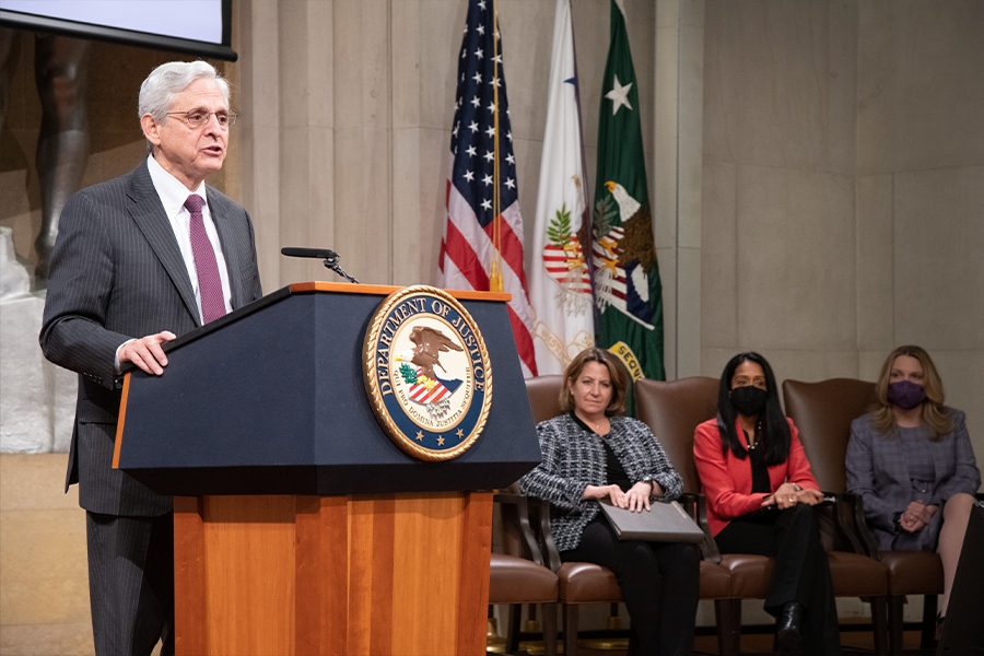 : Attorney General Merrick B. Garland speaks at a podium with the Department of Justice seal. To the right we see seated: Deputy Attorney General Lisa O. Monaco, Associate Attorney General Vanita Gupta, and Health and Human Services Deputy Secretary Andrea Palm.