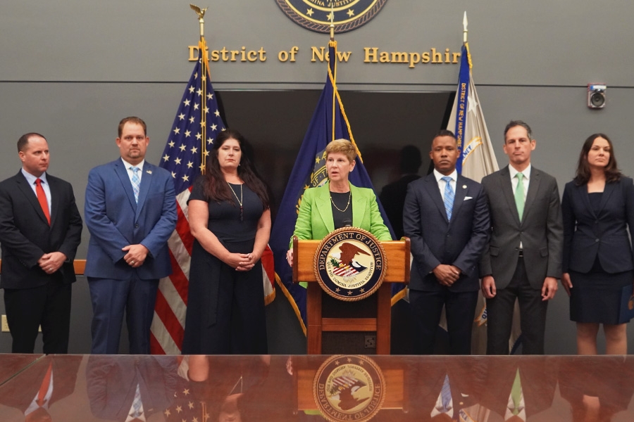 U.S. Attorney Jane E. Young for the District of New Hampshire speaks at the podium with a Department of Justice seal. The background displays the seal of the district of New Hampshire.