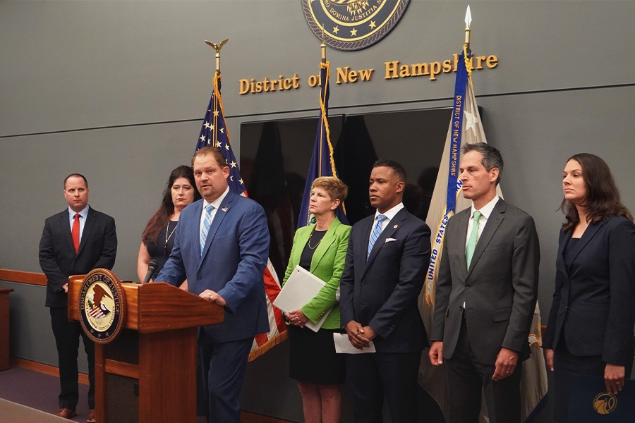 Deputy Inspector General Christian J. Schrank of the U.S. Department of Health and Human Services speaks at the podium with a Department of Justice seal. The background displays the seal of the district of New Hampshire.