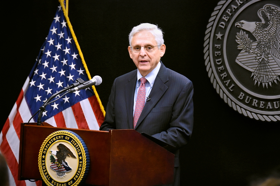 Attorney General Merrick B. Garland speaks at a podium bearing the Department of Justice seal. In the background is an American Flag and a Bureau of Prisons Seal.