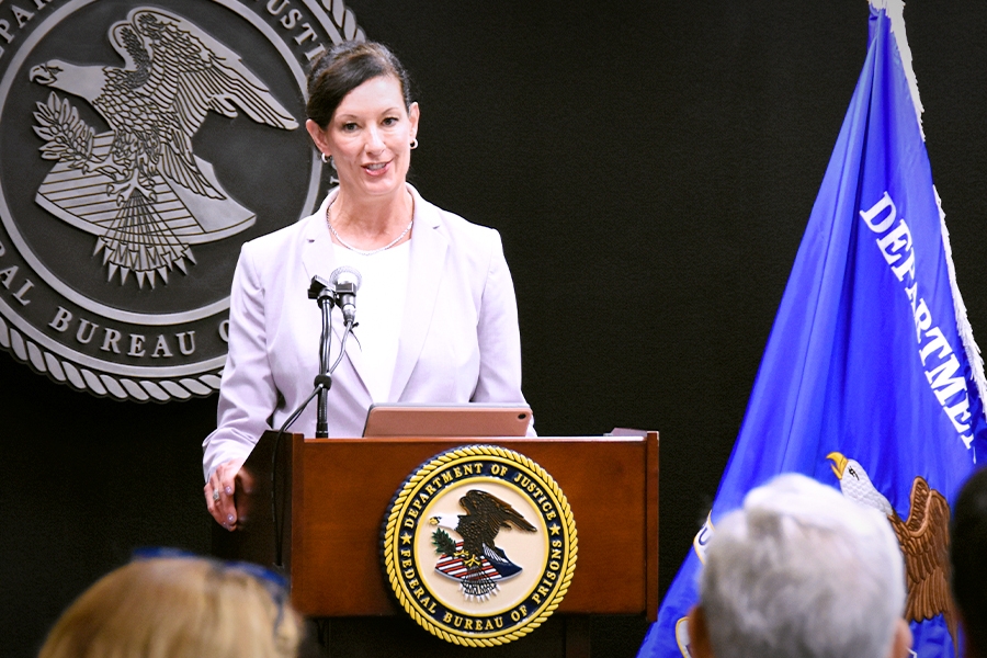 The Federal Bureau of Prisons Director Colette S. Peters speaks at a podium bearing the Department of Justice seal. In the background is the Bureau of Prisons seal. To the right is the blue Department of Justice flag.