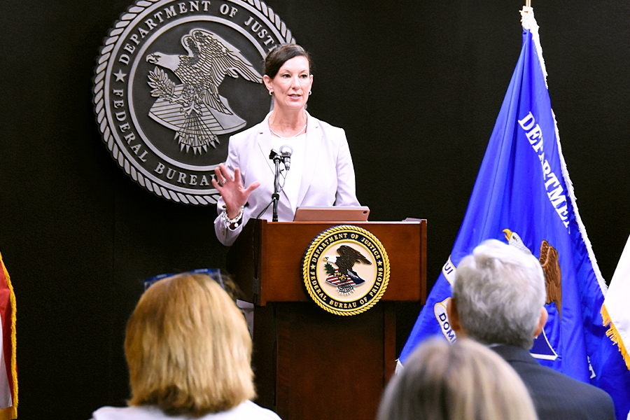 BOP Director Colette S. Peters speaks at a podium bearing the Department of Justice seal. In the background is the Bureau of Prisons seal. To the right is the blue Department of Justice flag.