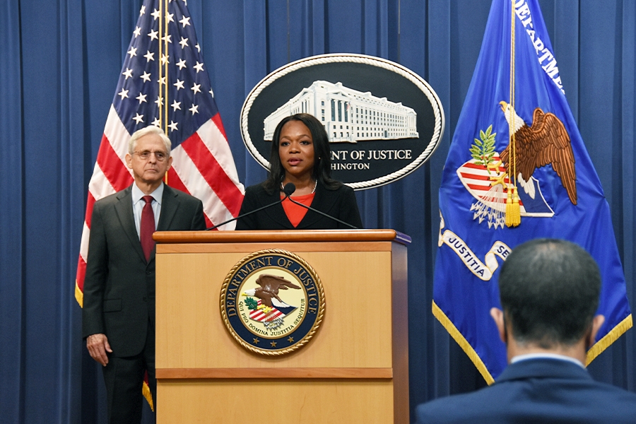 Assistant Attorney General Kristen Clarke speaks at a podium bearing the Department of Justice seal. To the left is Attorney General Merrick B. Garland, who stands in front of the American flag.