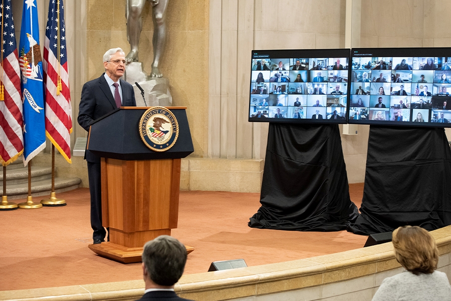 Attorney General Merrick Garland addresses the audience at the Department of Justice from a podium bearing the Department of Justice seal. To his right are screens showing virtual participants