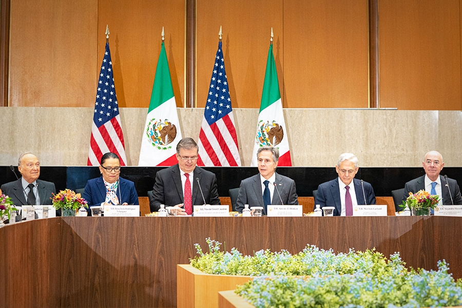 United States and Mexican Officials are seated in a conference room. The flags of the United States and Mexico are seen in the background