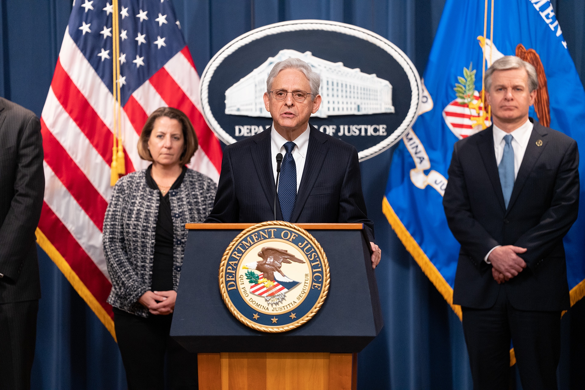 Attorney General Merrick B. Garland delivers remarks from a podium at the Department of Justice. Behind him stand FBI Director Christopher Wray (right) and Deputy Attorney General Lisa O. Monaco (left)