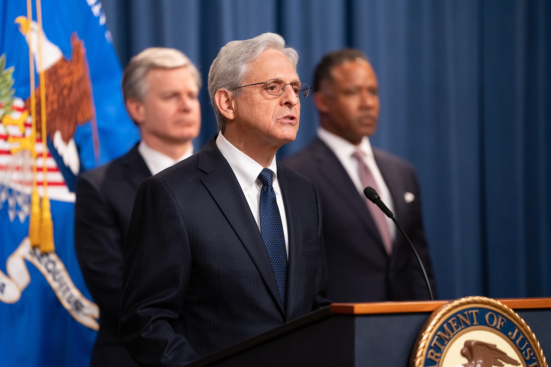 Attorney General Merrick B. Garland delivers remarks from a podium at the Department of Justice. Behind him stand FBI Director Christopher Wray (left) and Assistant Attorney General for the Criminal Division Kenneth A. Polite, Jr. (right)