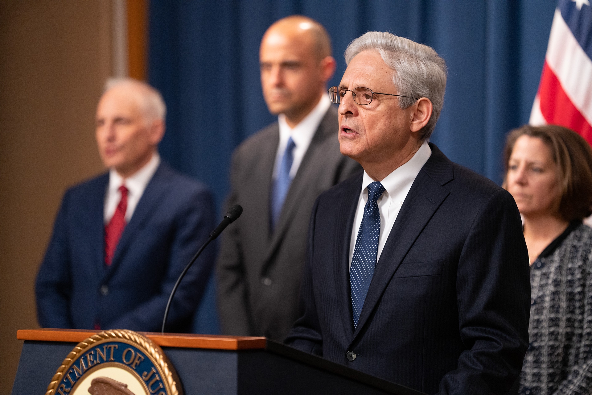 Attorney General Merrick B. Garland delivers remarks from a podium at the Department of Justice. Behind him stand Deputy Attorney General Lisa O. Monaco (right), U.S. Attorney for the District of Columbia Matthew M. Graves (left), and Assistant Attorney General for the National Security Division Matthew G. Olsen (far left)