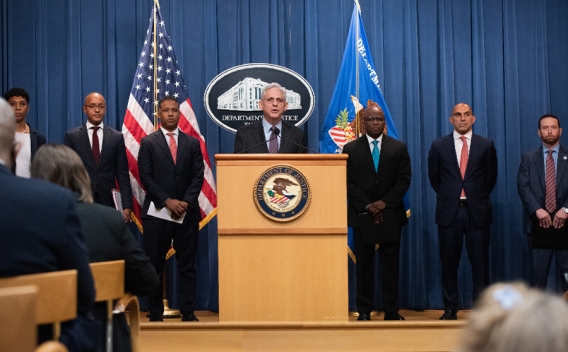 Attorney General Merrick B. Garland speaks from a podium to the crowd. Behind him, from left to right are Vanessa Roberts, Damian Williams, Kenneth A. Polite, Jr., Brian Turner, Luis Quesada, and Deputy Chief of the U.S. Postal Inspection Service Craig Goldberg.
