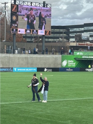 Two women stand on a soccer field. Their picture is on a big screen 