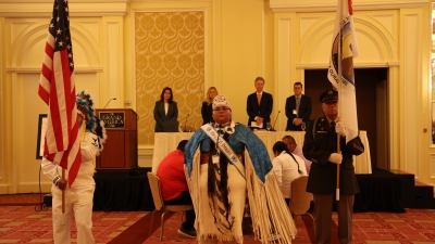 Four people standing behind a white table on stage. Ute Tribe drum group and Honor Guard/Presentation of colors in a ballroom 