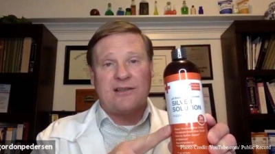 Alleged fake doctor in a white medical lab coat, in what appears to be an office, holding a bottle of "Silver Solution" and pointing to it in a screenshot from YouTube. Left bottom corner of picture says Gordon Pedersen. Right bottom corner of picture credit: YouTube/Public Record