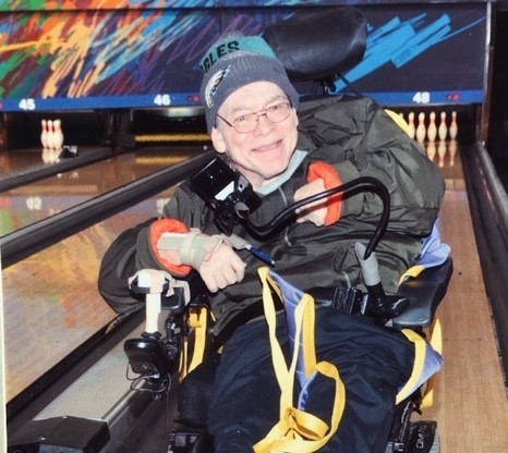 Thomas Graziani at a bowling alley, smiling, wearing a jacket and a beanie while in a wheelchair.. 