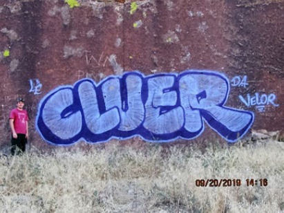 Defendant standing in front of "Cluer" and "Velor" graffiti on rock face at White River Narrows site in Lincoln County, Nevada. Person standing in front of Cluer and Velor graffiti on rock face.
