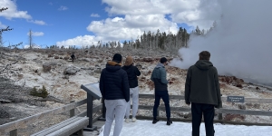 A man can be seen walking on thermal ground at the Steamboat Geyser in Yellowstone National Park. Four tourists look on from behind a fence on a wooden boardwalk. Image Credit: Yellowstone National Park
