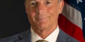 A smiling man with short light brown hair, wearing a blue suit with a white collared dress shirt and gold tie. He is standing facing the camera with a confident, relaxed smile, in front of a black background with an American Flag behind him. 