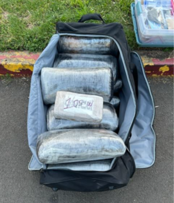 Law enforcement image. Black duffle bag filled with drugs wrapped in plastic and shaped into bricks. The bag sits on blacktop next to a red and yellow curb and grass. In the upper right is a plastic tote.