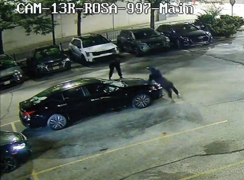 Video still of a camera footage of two people carjacking a black vehicle.