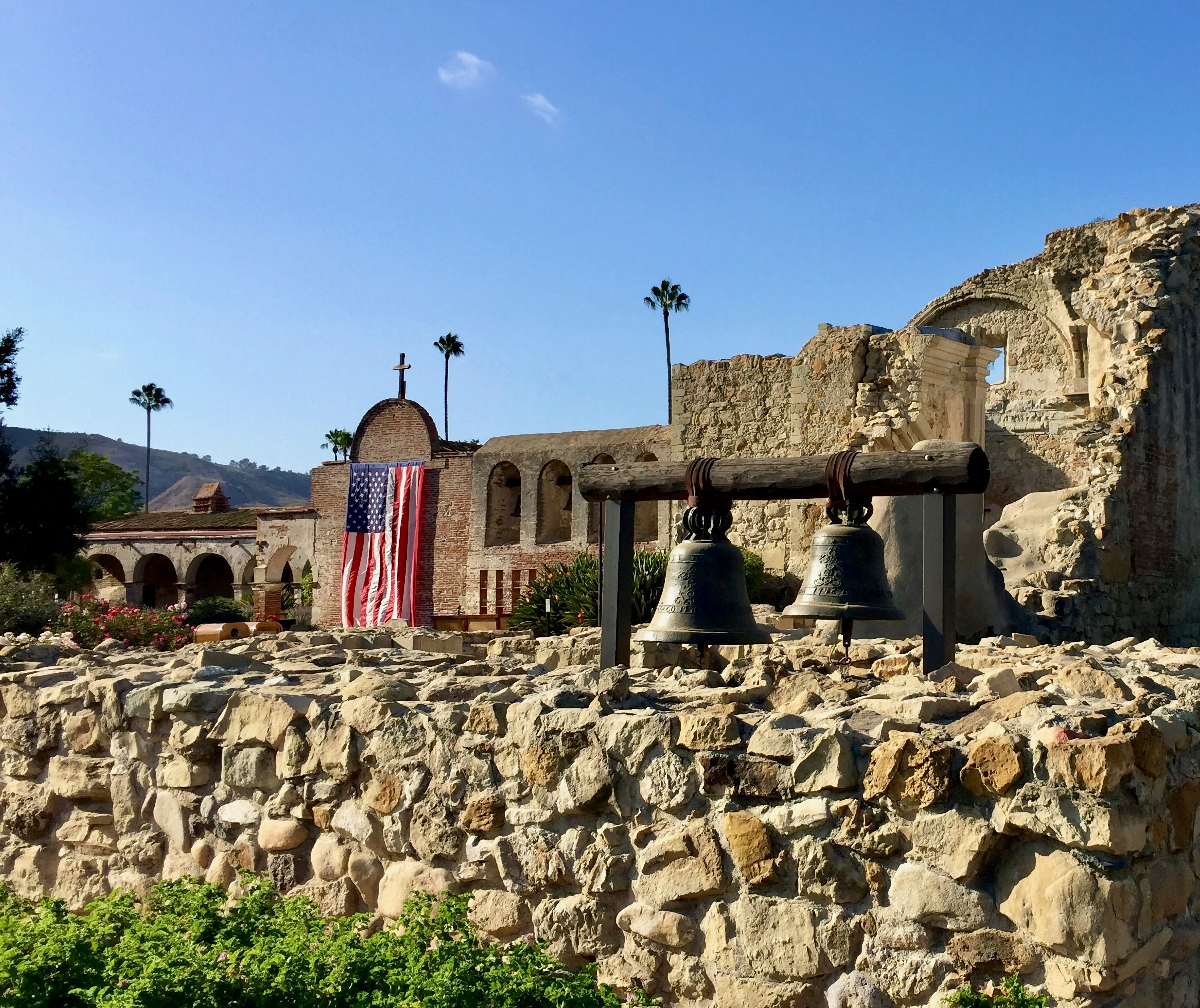 The ruins of the old church at California's Mission San Juan Capistrano with the U.S. flag draped near old church bells and palm trees in the background