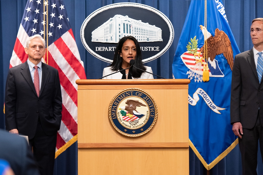 Associate Attorney General Vanita Gupta speaks at a podium bearing the Department of Justice seal. To the left is Attorney General Merrick B. Garland, who stands in front of the American flag. To the right stands the Principal Deputy Assistant Attorney for the Civil Division Brian Boynton.