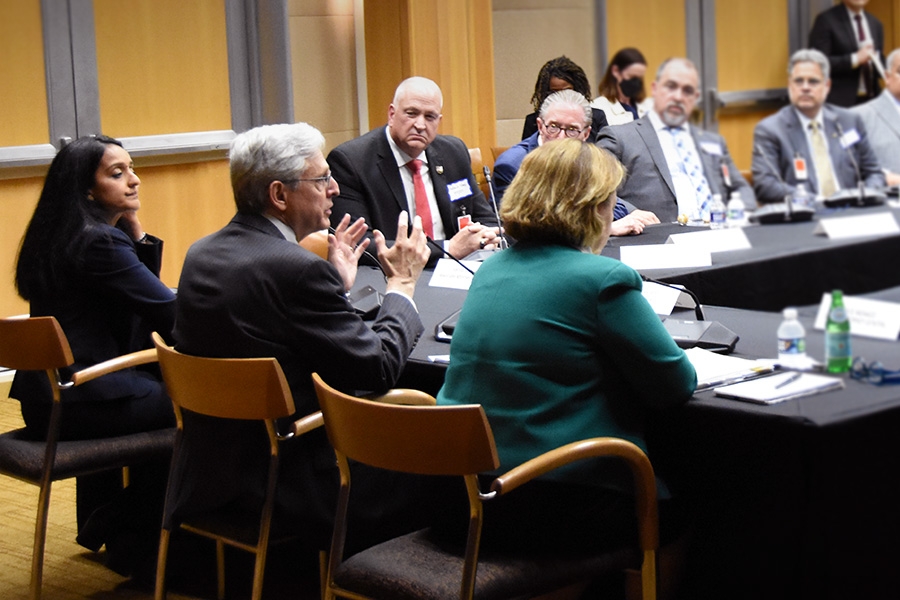 ASG Gupta, AG Garland, and DAG Monaco sit at a table. AG Garland addresses the crowd.