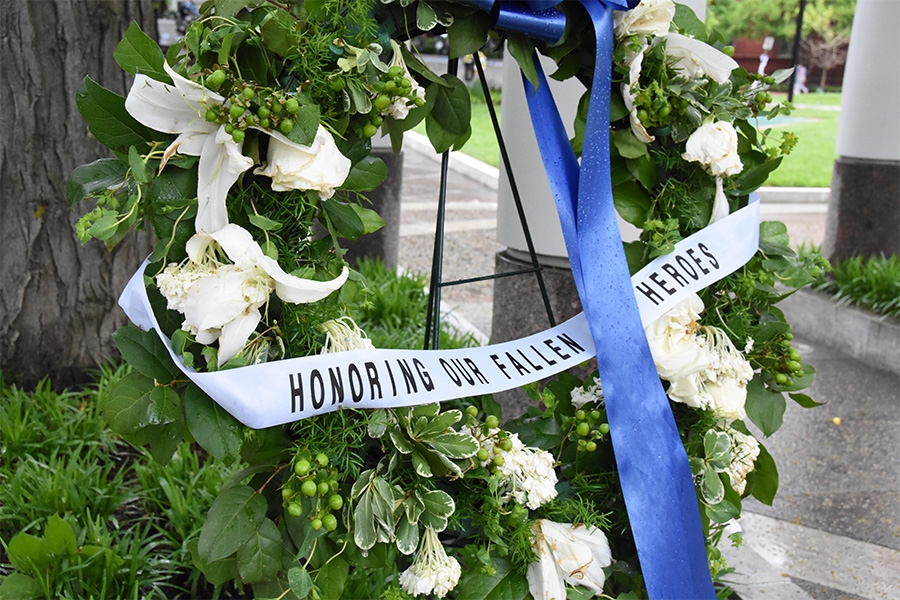 A wreath decorated with white lilies and a blue ribbon holds a sash that reads, “Honoring our fallen heroes”.