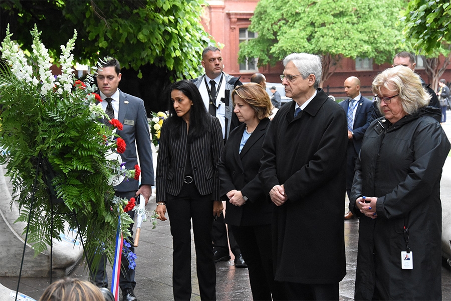 ASG Gupta, DAG Monaco, and AG Garland respectfully consider a wreath decorated with red, white, and blue flowers.
