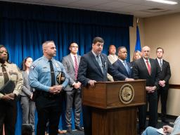 ATF Director Dettelbach delivers remarks from a podium. To the left stands Chief of the Minneapolis PD O'Hara (left) and Hennepin County Sheriff Witt (far left). To the right stands AAG Polite (right) and U.S. Attorney Luger (far right).