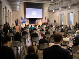 A crowd of officials are seated at the Department of Justice’s Great Hall. Merrick B. Garland stands at the podium.