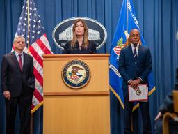 DEA Administrator Anne Milgram speaks at the podium. To the left is Attorney General Merrick B. Garland, and to the right is U.S. Attorney for the southern district of New York Damian Williams. An American flag, the Department of Justice seal, and the Department of Justice flag are seen in the background.