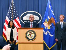 Attorney General Merrick B. Garland speaks at a podium bearing the Department of Justice seal. To the left is Associate Attorney General Vanita Gupta, who stands in front of the American flag. To the right stands the Principal Deputy Assistant Attorney for the Civil Division Brian Boynton.