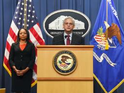 Attorney General Merrick B. Garland speaks at a podium bearing the Department of Justice seal. To the left is Assistant Attorney General Kristen Clarke, who stands in front of the American flag.