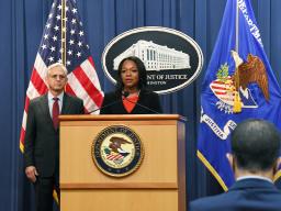 Assistant Attorney General Kristen Clarke speaks at a podium bearing the Department of Justice seal. To the left is Attorney General Merrick B. Garland, who stands in front of the American flag.