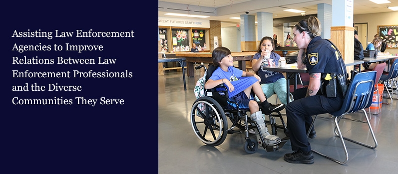 A female police officer engaging with a young boy in a wheelchair and a young girl eating lunch at a school.