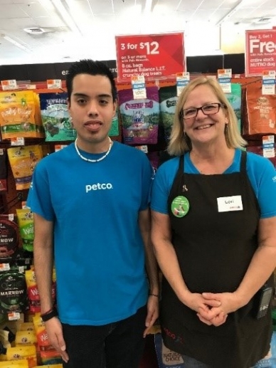 Genaro is pictured in front of a store shelf standing next to a coworker. Both are wearing blue shirts. 
