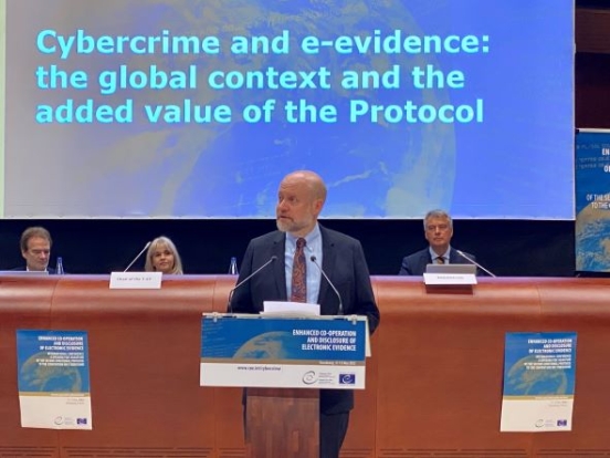 DAAG Richard Downing hablando en la Convención de Budapest Hombre parado detrás de un podio hablando con una audiencia con una pantalla grande a la vista